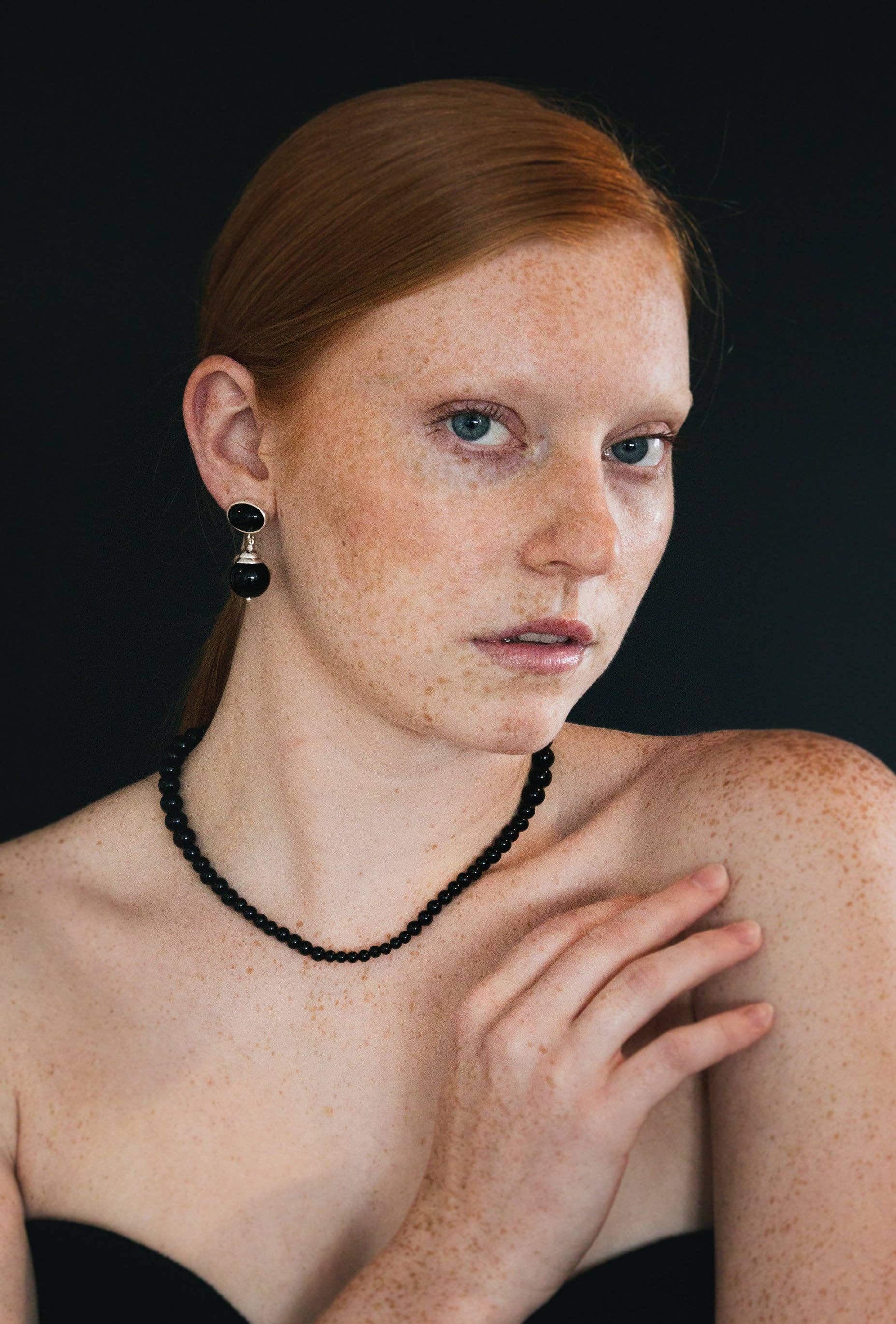 Woman with red hair and freckles wearing a black beaded necklace against a black background