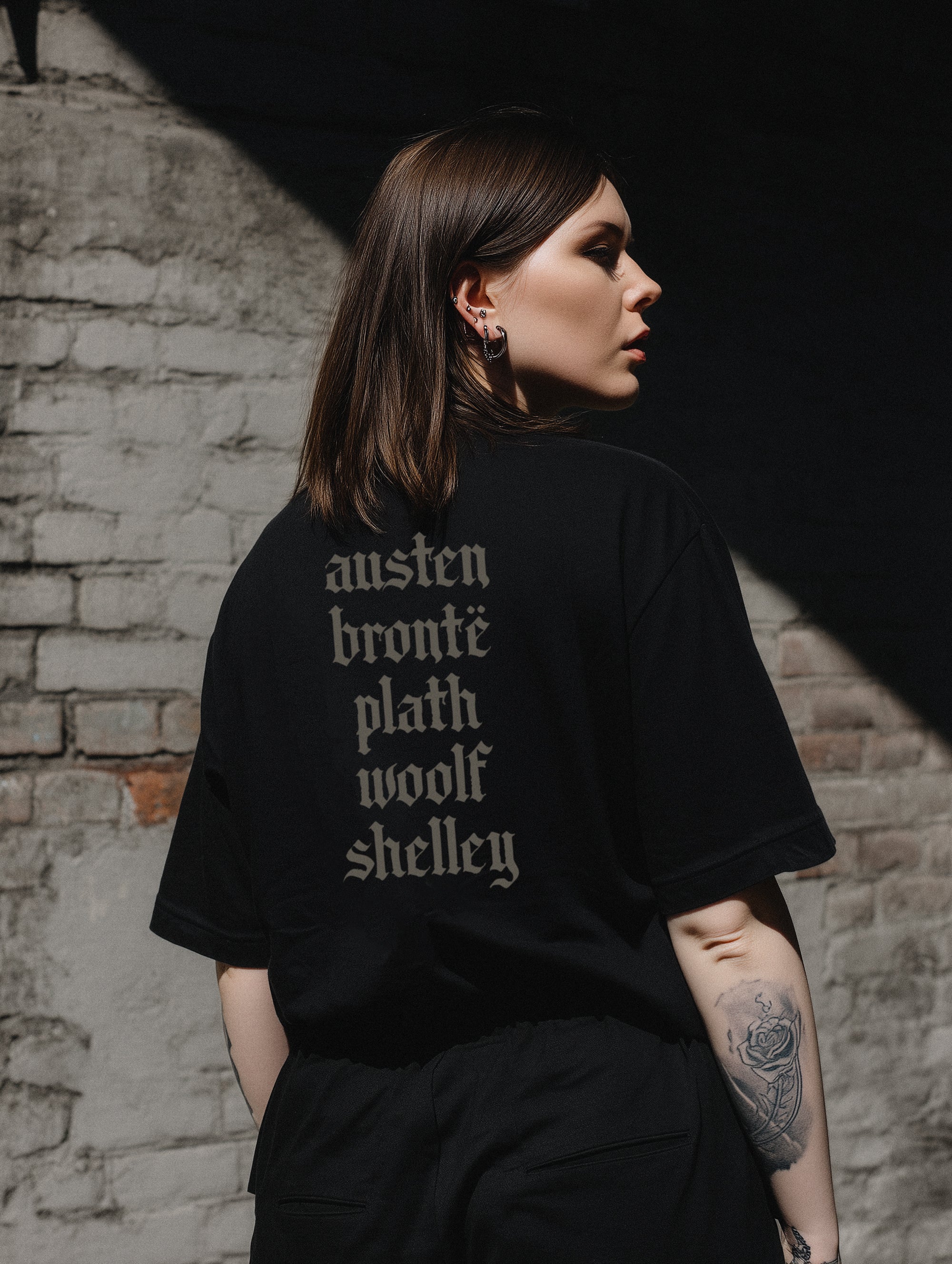 A person wearing a white crew neck t-shirt with text on the back. The text is a list of names: austen, bronte, plath, woolf, shelley.