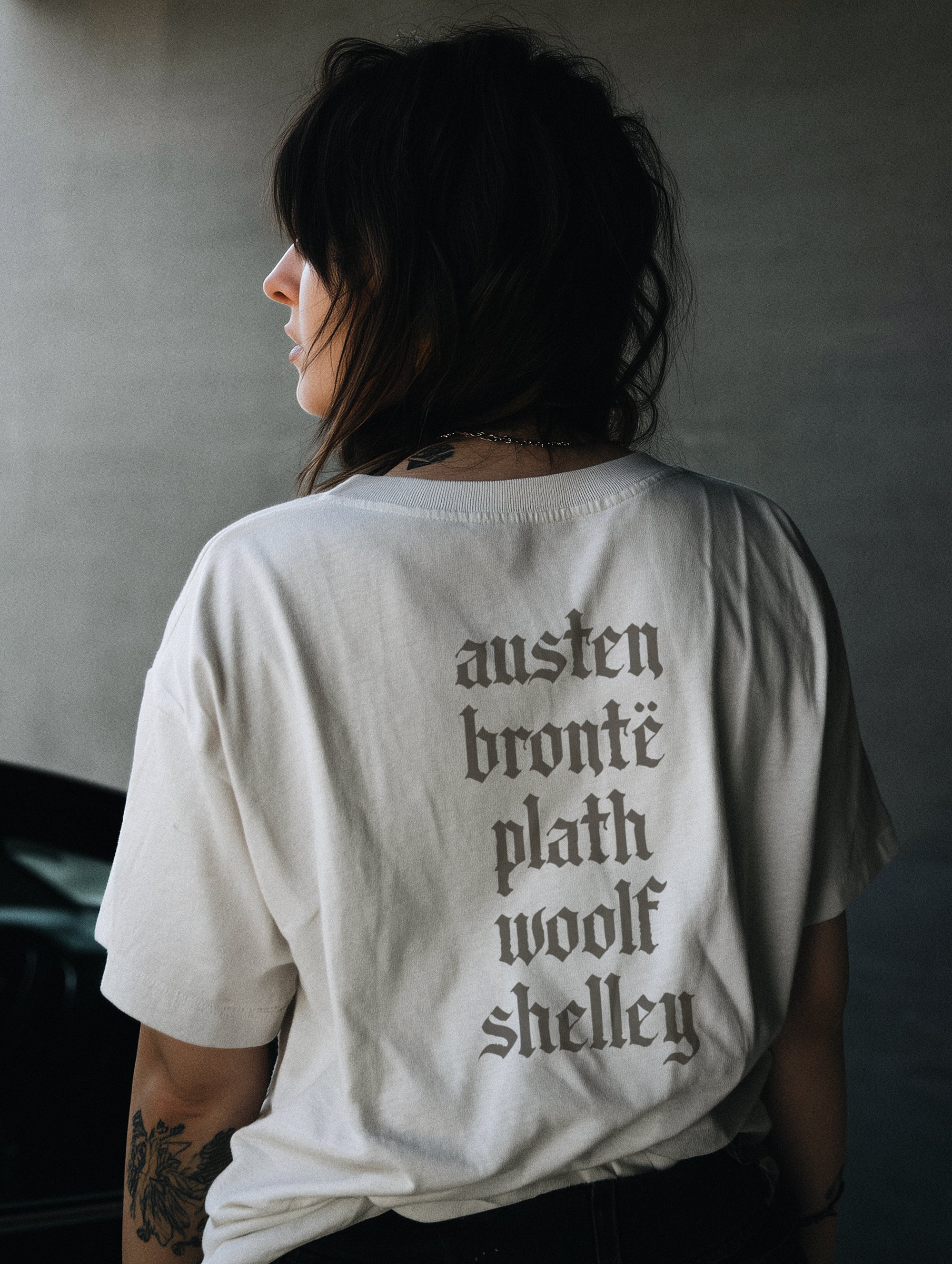 A person wearing a white crew neck t-shirt with text on the back. The text is a list of names: austen, bronte, plath, woolf, shelley.