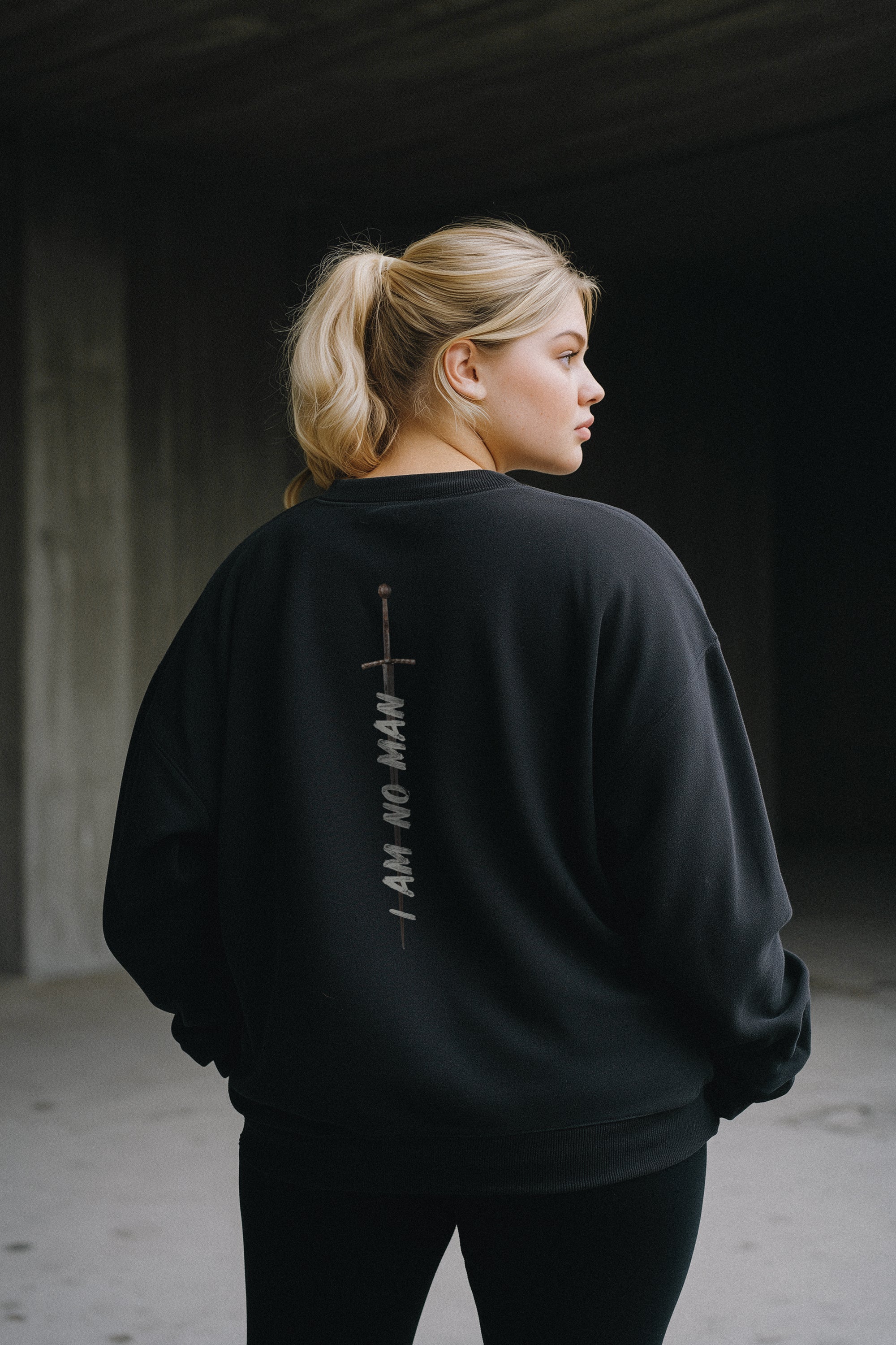 Woman wearing a black sweatshirt with "I Am No Man" and a sword graphic on the back, standing against a dark background.