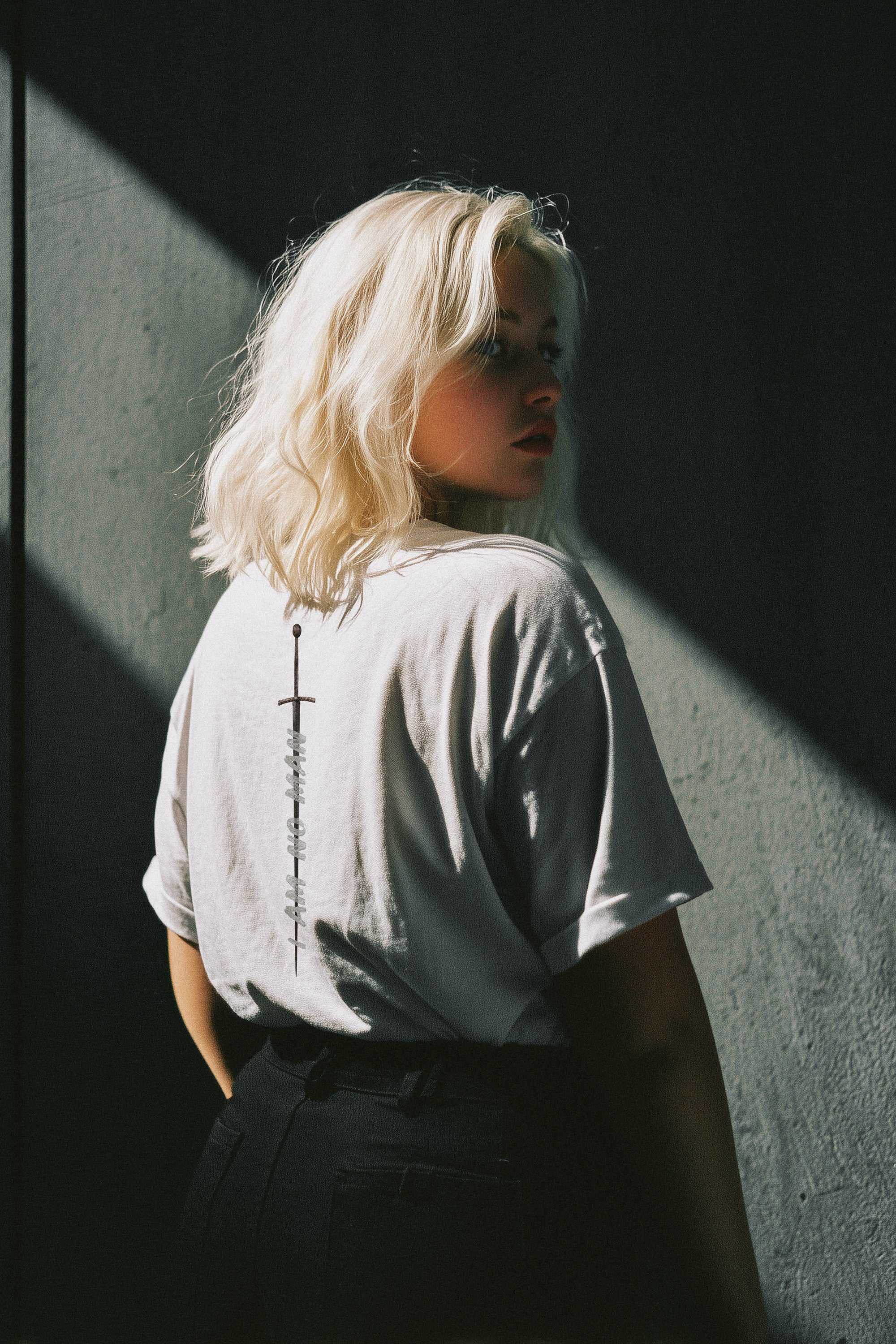 Person with blonde hair wearing a light-colored shirt against a dark wall.