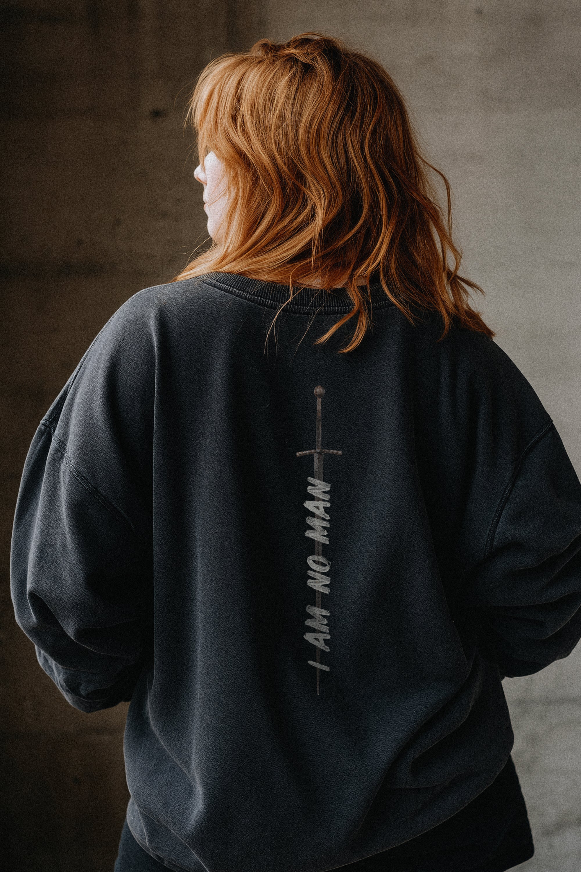 Woman wearing a black sweatshirt with "I Am No Man" and a sword graphic on the back, standing against a dark background.