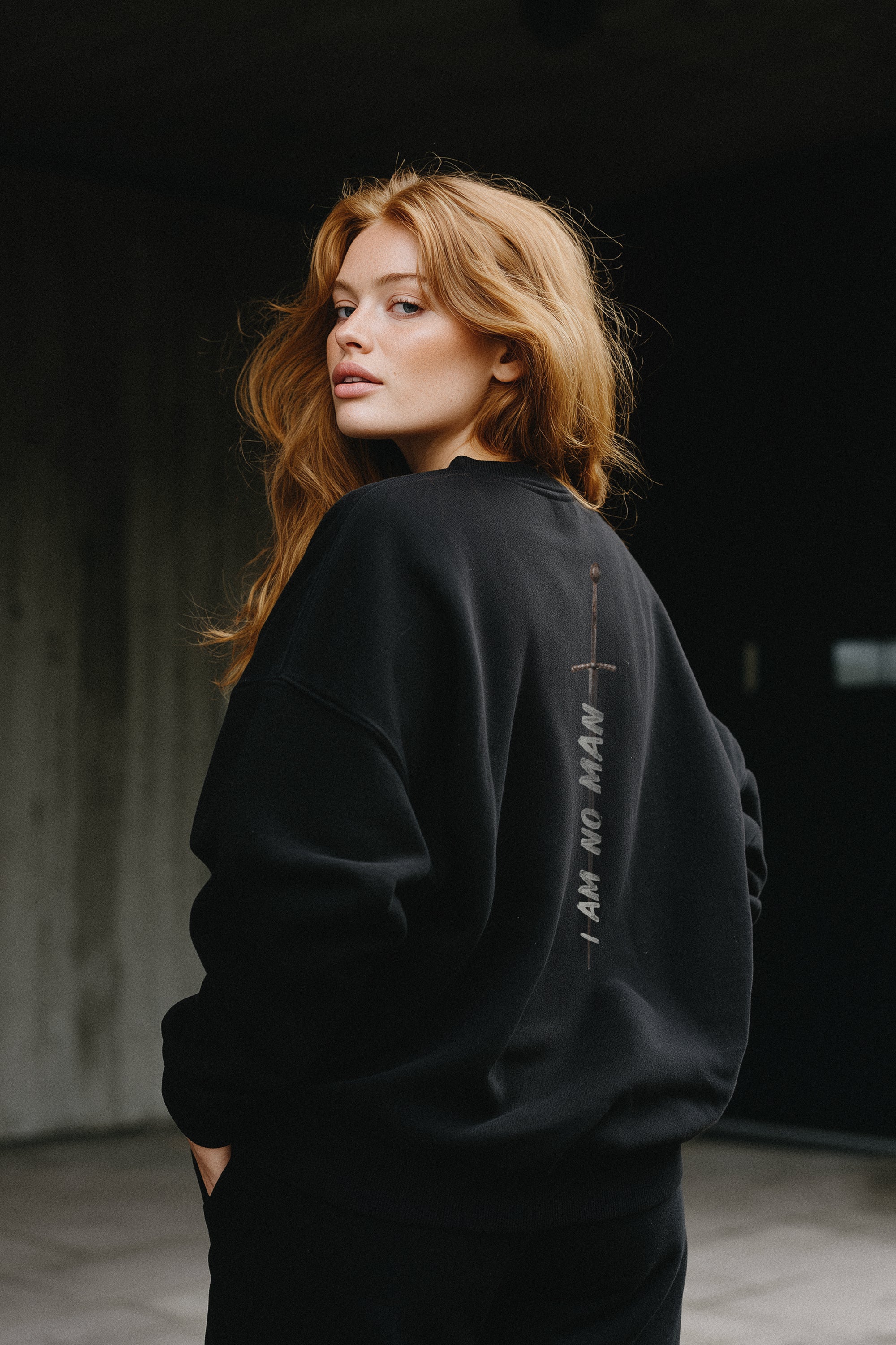 Woman wearing a black sweatshirt with "I Am No Man" and a sword graphic on the back, standing against a dark background.